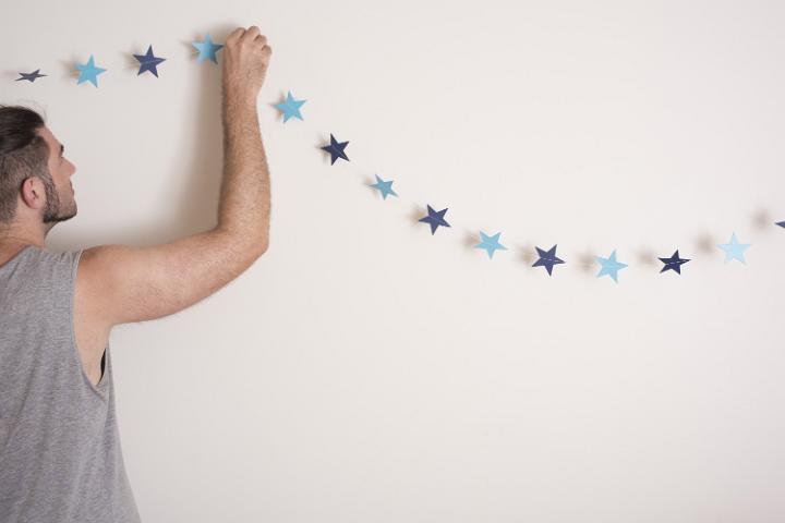 Man hanging Christmas decorations at home attaching a garland of festive blue stars to a white wall with copy space