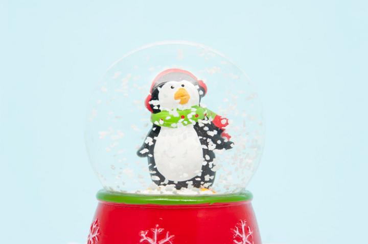 Cute little penguin in a winter scarf and cap standing in swirling snowflakes in a Christmas snow globe decoration against a light blue background with copyspace