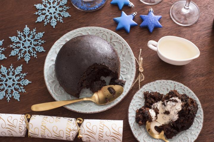 Serving of traditional fruity Christmas pudding with brandy sauce or cream on a festive decorated table with the whole dessert and served portion plated side by side