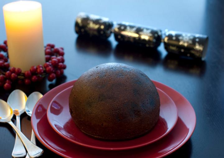 Large traditional steamed spiced Christmas fruit pudding served on festive red plates on the dinner table with a glowing candle and berry wreath to celebrate the Xmas season