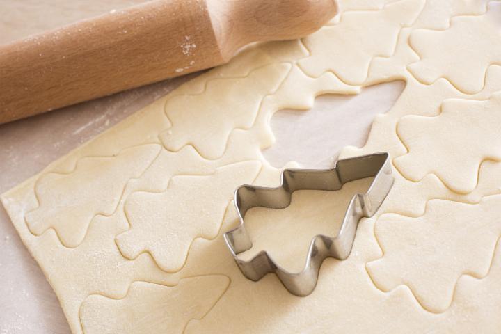 Making Christmas tree cookies in a home kitchen with rolled uncooked pastry with cut out shapes and a metal cookie cutter alongside a wooden rolling pin
