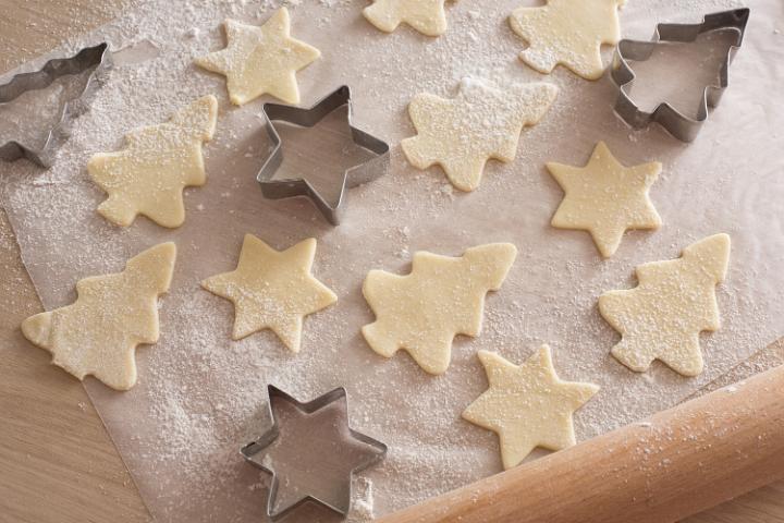 Making traditional Christmas cookies at home with star and tree shaped cookie cutters alongside cut out uncooked dough on floured paper on the kitchen counter