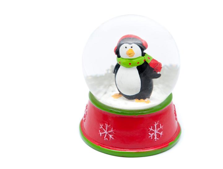Cute little penguin in scarf and knitted winter hat standing in a Christmas snow globe standing in the snow waiting to be shaken to activate the snowflakes for a snowstorm effect, isolated on white